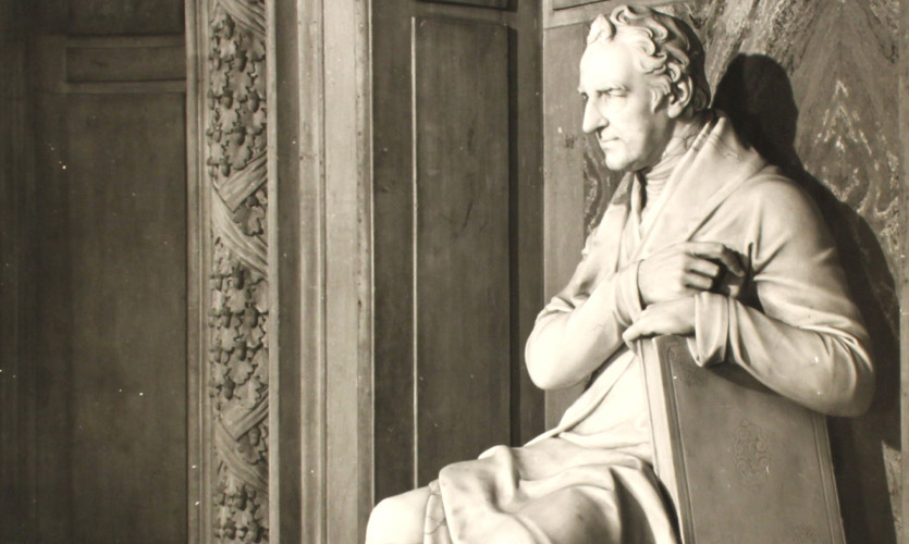 A marble statue of William Roscoe, he has been sculpted sitting down with his arms clasped around a book