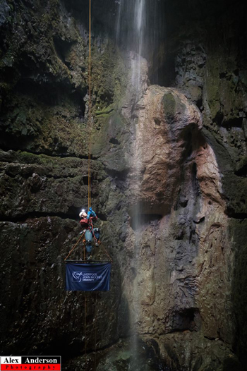 Jack Overhill climbing out of Alum Pot, a pothole with waterfall in Yorkshire, holding an LJMU flag