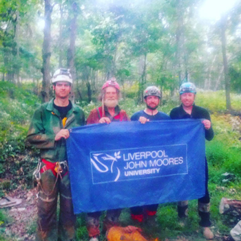 Four members of the Germany Valley Karst Survey team holding an LJMU flag in the forest