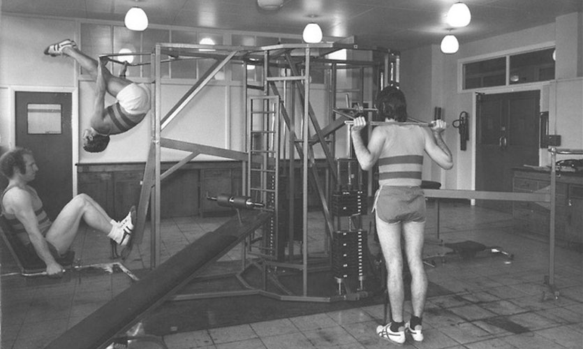 Black and white photo of three men exercising in a gym in 1974