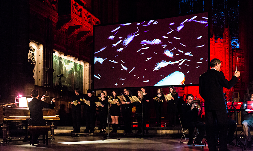 A choir being lead in front of a screen with a series of lights projected onto it