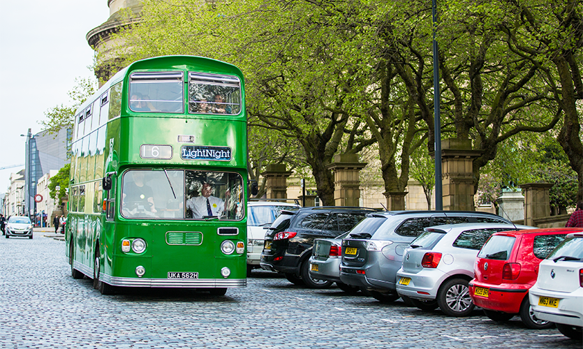A green bus going to "LightNight" driving down a leafy cobbled street