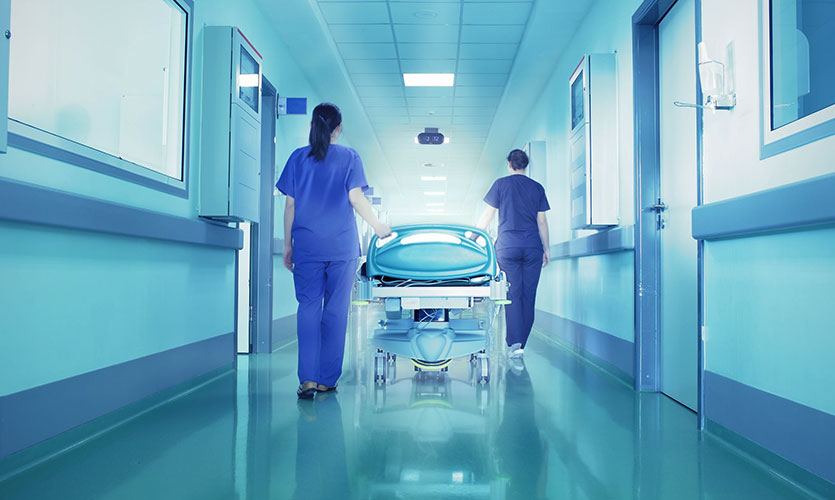 Image of two nurses pushing a hospital bed through a corridor towards a bright light.