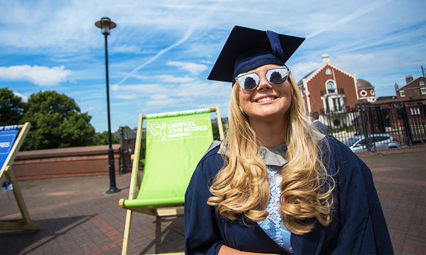 Smiling girl dressed in a gown and mortar board wearing sunglasses that are reflecting the Liverpool Cathedral