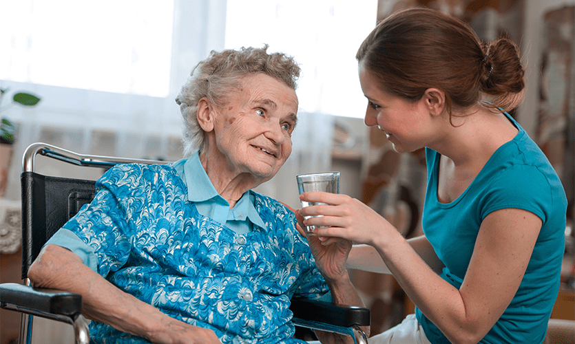 Image of a frail old woman in a wheelchair being given a drink by a carer.