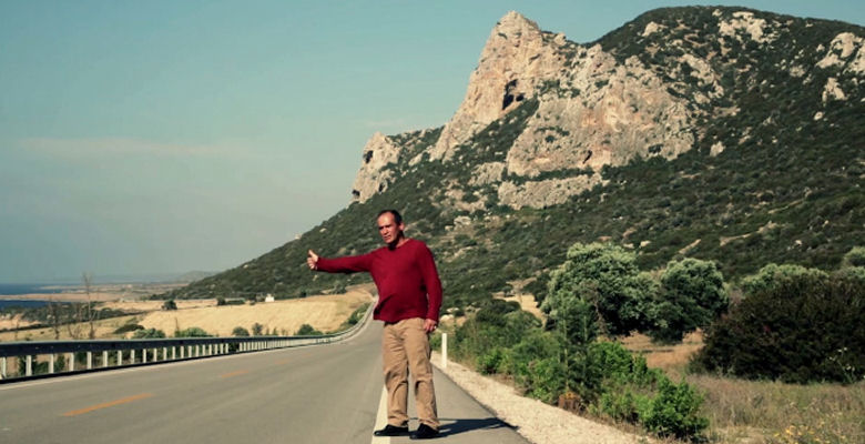Image of a film still of man standing in road between mountains hitching a lift