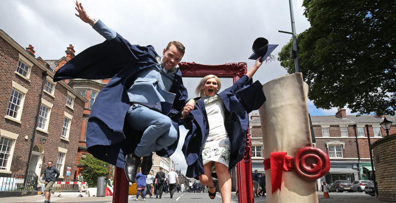 Image of two graduates in cape and gown jumping through a picture frame on Hope Street