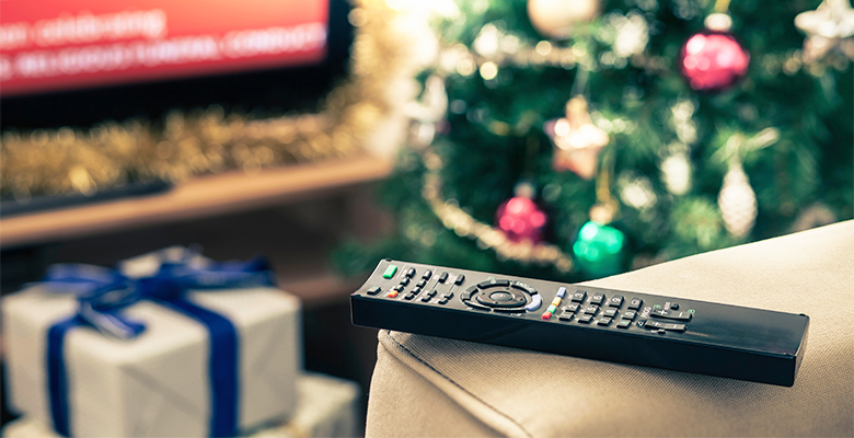 Image of a remote control resting on a sofa with a backdrop of a Christmas tree with presents under it.