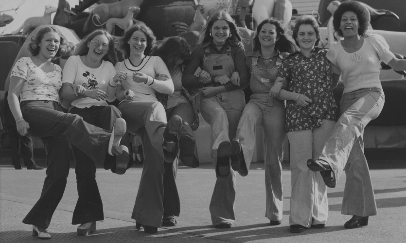 A black and white photograph of a group of seven female Littlewoods workers each with one leg kicked up in the air as they pose for the photo on a day out in Blackpool