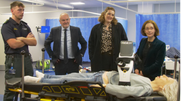 A paramedic student stood with VC Professor Mark Power, Stephanie Harris and Vivienne Stern from Universities UK, they are all standing behind a medical manikin lying on a hospital bed 