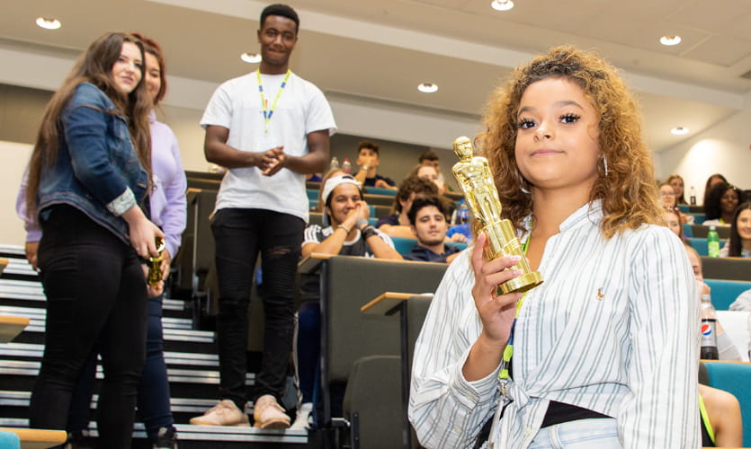 Photo of a teenage girl holding an oscar statue and smiling, in a lecture hall with other students