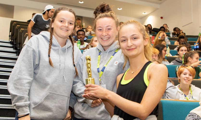 Three teenagers holding a plastic oscar statue in a lecture theatre smiling at the camera at the LJMU summer school