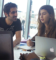 Two students sitting at a table with laptops on it in conversation.