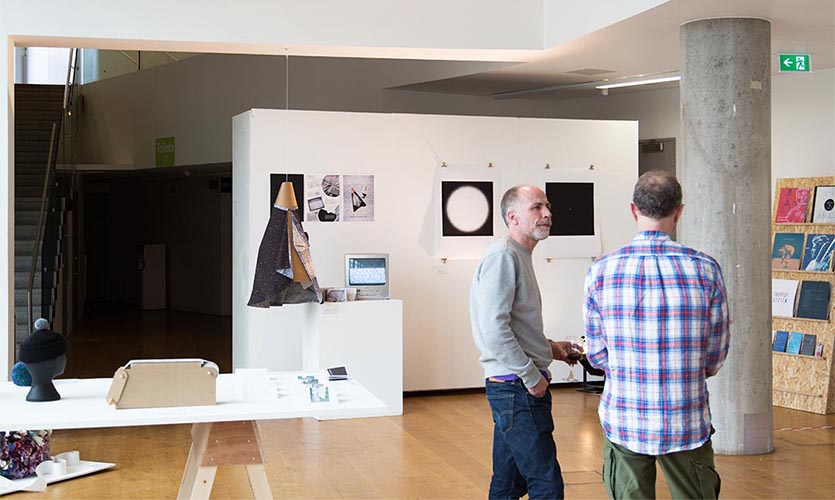 Image of two men in conversation in the exhibition space with paintings on a wall in the background.